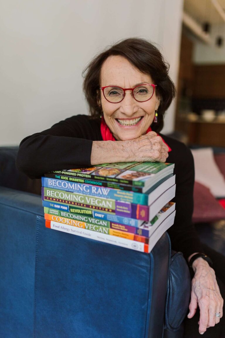 An older woman with glasses and a warm smile leans on a stack of books on a blue sofa, their spines revealing her passion for vegan themes. She wears a black top and red scarf, adding to the cozy ambiance of the room with its softly blurred background.