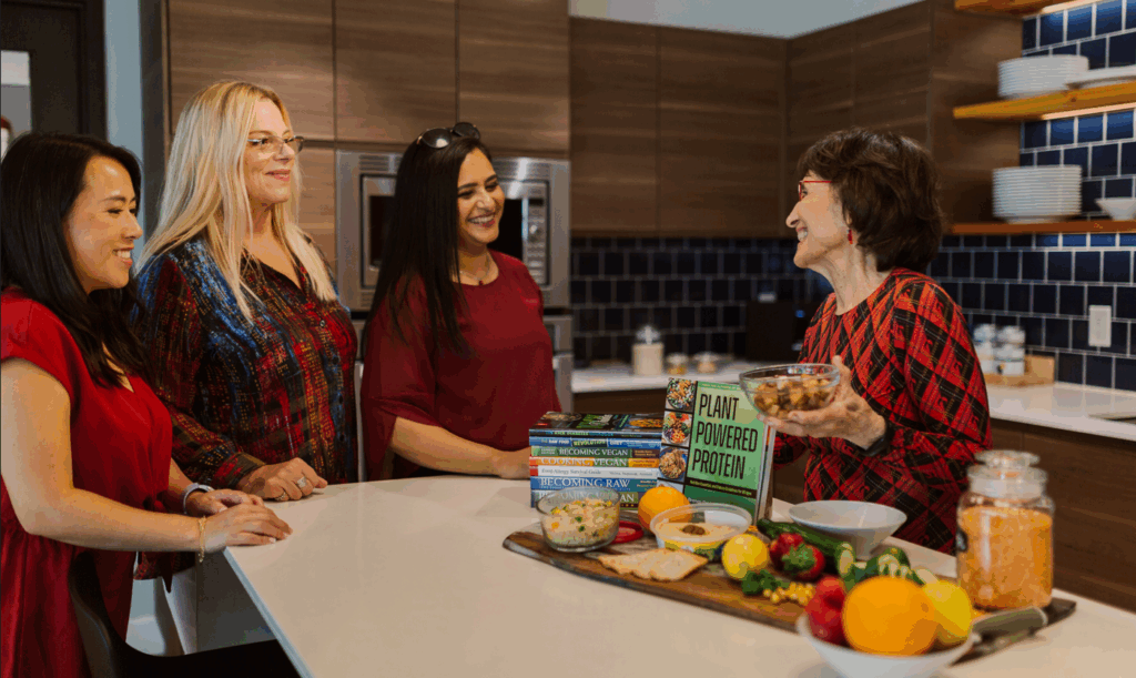 Four women stand in a kitchen, engaged in lively conversation, perhaps inspired by their favorite podcasts on plant-based cooking. A countertop displays cookbooks and ingredients like vegetables and lentils. Wooden cabinets and blue tiled walls provide a warm backdrop to their culinary gathering.