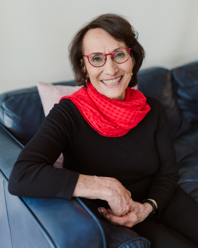 A smiling person with short dark hair and red glasses sits on a blue sofa, reviewing their resumé. They wear a black top and a red scarf.