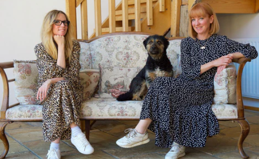 Two women are sitting on a floral-patterned sofa with a black dog between them. The woman on the left wears a leopard print dress and glasses, while the woman on the right wears a polka dot dress. Both are wearing white sneakers. A staircase is visible behind them.