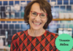 Smiling woman with short brown hair and red glasses, wearing a red and black patterned shirt. She stands in front of a blue tiled wall. A green label in the corner reads Venassa Maison.
