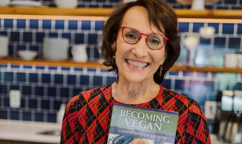 A person with short dark hair and red glasses is smiling and holding a book titled Becoming Vegan: Comprehensive Edition. They are wearing a red and black patterned top, and there are shelves with kitchen items in the background.